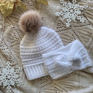 May include: White crocheted beanie with a fluffy brown pom pom and a matching white crocheted headband. The beanie and headband are laid out on a beige crocheted fabric.