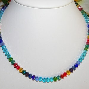 May include: A colorful beaded necklace displayed on a white bust. The necklace features a gradient of colors, including blue, green, yellow, and red. The beads are small and faceted, catching the light.