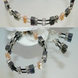 May include: A black and silver necklace and earrings set with square-shaped hematite beads and clear crystal accents. The necklace and earrings feature a double strand design with peach-colored beads.