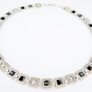 May include: Silver-toned necklace featuring a repeating pattern of square links. Each square contains a dark bead, with smaller beads connecting the squares. The necklace is displayed on a white background.