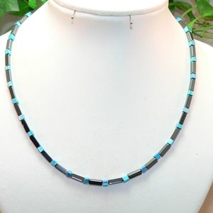 Può includere: Una collana fatta di perline quadrate di colore grigio scuro e blu chiaro. La collana è su un manichino bianco.