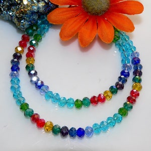 May include: A multi-strand necklace featuring faceted beads in a rainbow of colors. The beads transition from red to yellow, green, blue, and purple. An orange flower and a cluster of blue and gold crystals are in the background.