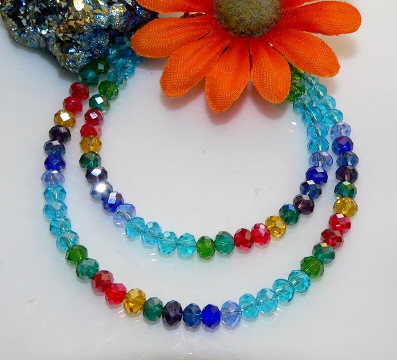 May include: A multi-strand necklace featuring faceted beads in a rainbow of colors. The beads transition from red to yellow, green, blue, and purple. An orange flower and a cluster of blue and gold crystals are in the background.