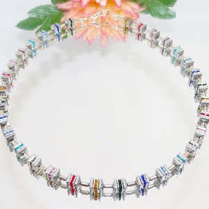 Puede incluir: Un collar plateado con cuentas cuadradas de colores. Cada cuenta está adornada con múltiples cristales pequeños de varios colores, creando un efecto arcoíris.