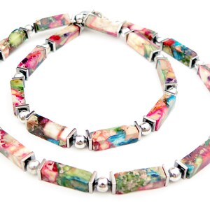 Puede incluir: Un collar colorido con cuentas rectangulares en tonos rosa, verde, azul y crema, conectadas por detalles metálicos plateados. El collar se muestra sobre un fondo blanco.