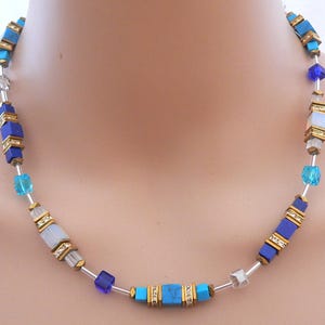 Könnte beinhalten: Halskette mit blauen und türkisfarbenen Würfelperlen, goldfarbenen Akzenten und klaren Kristallperlen. Die Perlen sind auf einem silberfarbenen Draht aufgefädelt. Das Design der Halskette umfasst verschiedene Blautöne.