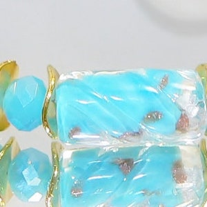 May include: A close-up of a turquoise beaded bracelet with gold-colored accents. The bracelet features cylindrical beads with a marbled effect, interspersed with gold-colored metal rings and faceted turquoise beads. The bracelet is displayed on a reflective surface.