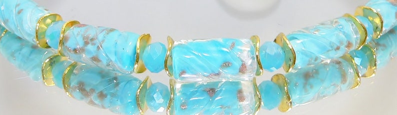 May include: A close-up of a turquoise beaded bracelet with gold-colored accents. The bracelet features cylindrical beads with a marbled effect, interspersed with gold-colored metal rings and faceted turquoise beads. The bracelet is displayed on a reflective surface.