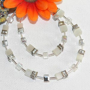 May include: A necklace with a delicate design. The necklace features alternating square and rectangular beads in clear and off-white colors, with silver-toned metal accents. The beads are strung together, creating a layered effect. The necklace is a piece of jewelry.