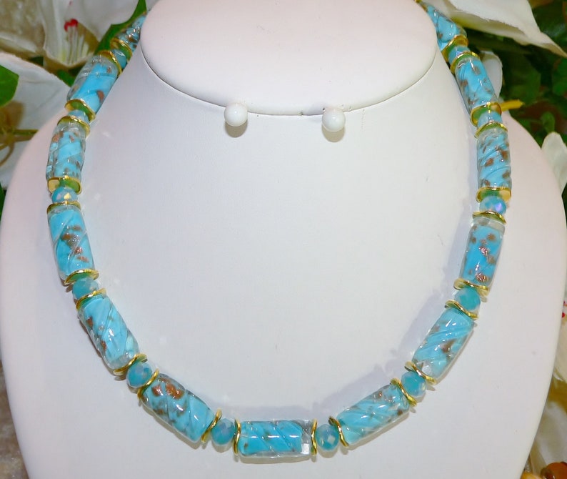 May include: A turquoise beaded necklace with gold accents. The necklace features cylindrical beads with a swirled pattern and gold-colored spacers. The necklace is displayed on a white bust, showcasing the jewelry's design and color.