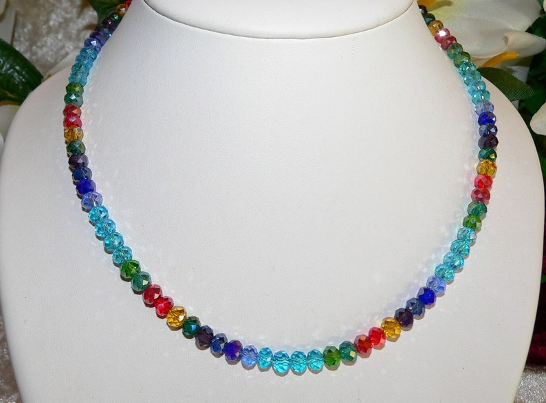 May include: A colorful beaded necklace displayed on a white bust. The necklace features a gradient of colors, including blue, green, yellow, and red. The beads are small and faceted, catching the light.