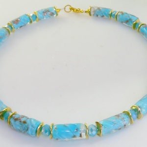 May include: A turquoise glass bead necklace with gold-colored accents. The necklace features cylindrical beads with a swirled pattern, separated by small gold-colored rings. The clasp is gold-colored.