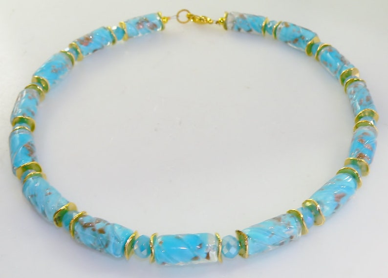 May include: A turquoise glass bead necklace with gold-colored accents. The necklace features cylindrical beads with a swirled pattern, separated by small gold-colored rings. The clasp is gold-colored.