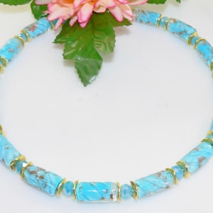 May include: A turquoise beaded necklace with gold accents. The necklace features cylindrical beads with a swirled pattern and gold-colored spacers. The necklace is displayed on a white surface, with a pink and green floral arrangement in the background.