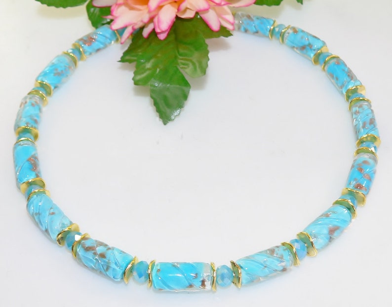 May include: A turquoise beaded necklace with gold accents. The necklace features cylindrical beads with a swirled pattern and gold-colored spacers. The necklace is displayed on a white surface, with a pink and green floral arrangement in the background.