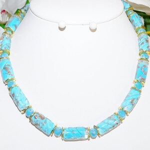 May include: A turquoise beaded necklace with gold accents. The necklace features cylindrical beads with a swirl pattern and gold-colored metal spacers. The matching earrings are visible in the background. The jewelry is displayed on a white mannequin.