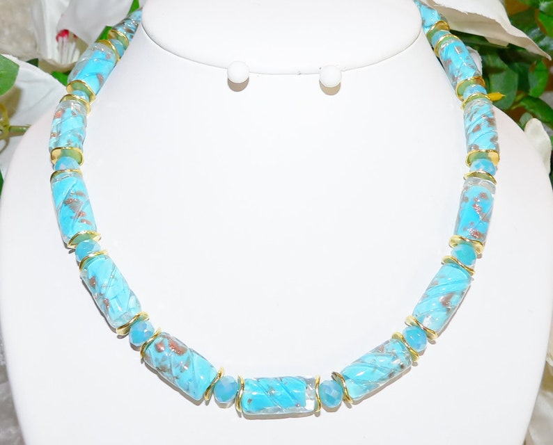 May include: A turquoise beaded necklace with gold accents. The necklace features cylindrical beads with a swirl pattern and gold-colored metal spacers. The matching earrings are visible in the background. The jewelry is displayed on a white mannequin.