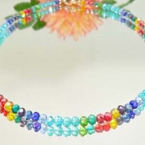 May include: A multi-colored beaded necklace with a rainbow of colors, including red, yellow, green, blue, and turquoise. The necklace is made of small, faceted beads and is displayed on a reflective surface.