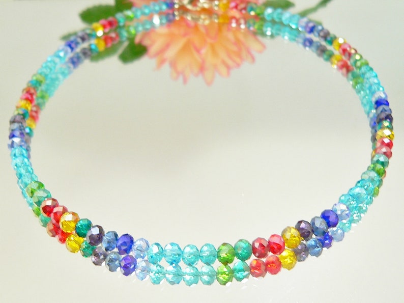 May include: A multi-colored beaded necklace with a rainbow of colors, including red, yellow, green, blue, and turquoise. The necklace is made of small, faceted beads and is displayed on a reflective surface.