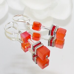 May include: A pair of dangle earrings featuring orange and red cube-shaped beads. The earrings have silver-colored metal accents and lever-back closures. The beads are arranged in a stacked design.
