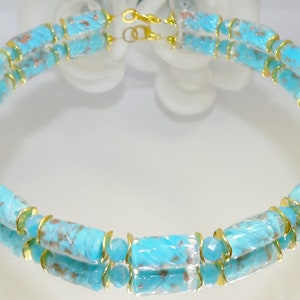May include: A turquoise necklace with gold-colored accents. The necklace features cylindrical beads with a carved design and gold-colored metal rings. The clasp is gold-colored. The necklace is displayed on a reflective surface.