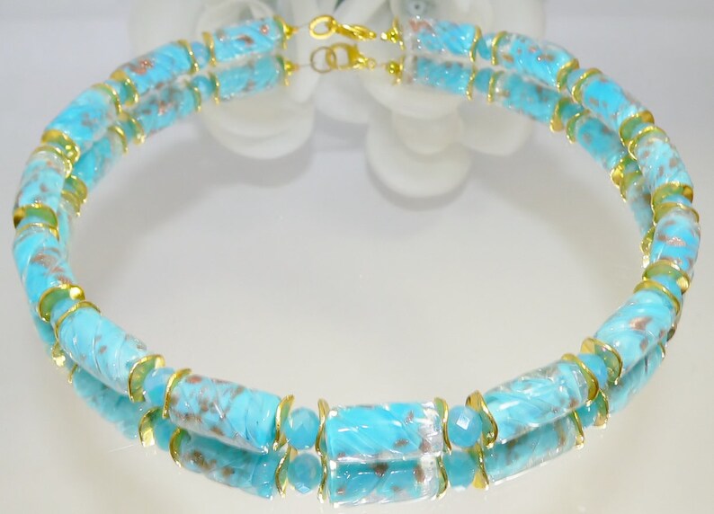 May include: A turquoise necklace with gold-colored accents. The necklace features cylindrical beads with a carved design and gold-colored metal rings. The clasp is gold-colored. The necklace is displayed on a reflective surface.