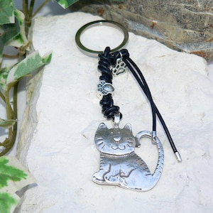 May include: A silver cat charm keychain with a black leather cord and silver paw print beads. The charm has a heart-shaped tail and a smiling face.