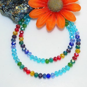 May include: Two strands of colorful beaded necklaces. The beads are arranged in a rainbow pattern, transitioning from red to blue. An orange flower and a piece of dark, textured stone are in the background.
