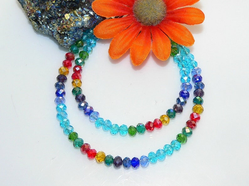 May include: Two strands of colorful beaded necklaces. The beads are arranged in a rainbow pattern, transitioning from red to blue. An orange flower and a piece of dark, textured stone are in the background.