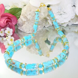 May include: A turquoise beaded necklace with gold-colored accents. The necklace features cylindrical and faceted beads in shades of blue and brown, connected by gold-toned metal. The necklace is displayed on a reflective surface, with flowers in the background.
