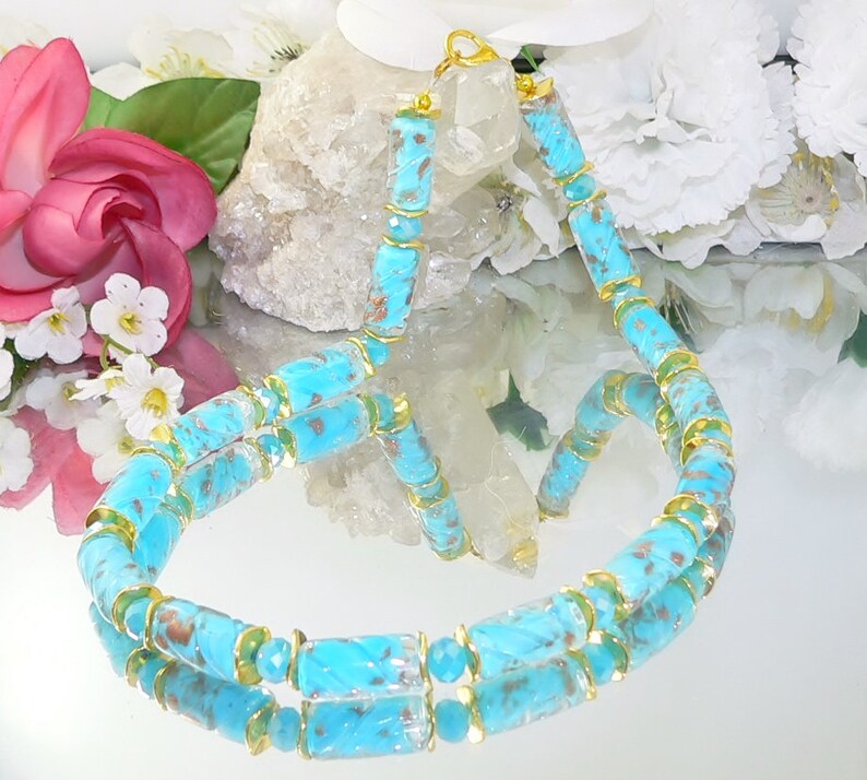 May include: A turquoise beaded necklace with gold-colored accents. The necklace features cylindrical and faceted beads in shades of blue and brown, connected by gold-toned metal. The necklace is displayed on a reflective surface, with flowers in the background.