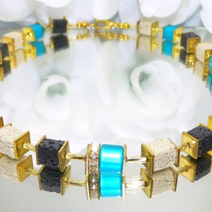 May include: A gold necklace with black, white, and turquoise square beads. The beads are made of lava rock, white stone, and glass.