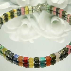 May include: A colorful necklace made of square-shaped gemstone beads. The beads are arranged in a rainbow pattern, with each bead a different color. The necklace is made of silver metal and has a clasp.