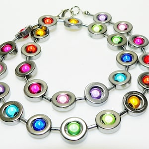 Peut inclure: Un collier en argent avec des perles rondes argentées. Chaque perle a une plus petite perle colorée au centre. Les couleurs des perles sont le rouge, l'orange, le jaune, le vert, le bleu, le violet et le rose.