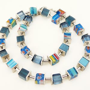 May include: A necklace featuring square, colorful glass beads in shades of blue, red, and yellow, connected by silver-toned metal links. The beads have various patterns and designs. The necklace is displayed on a white background.