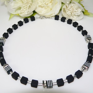 May include: A black and white necklace with square beads and silver accents. The necklace is made of black beads with white stripes and silver accents. The necklace is on a white background.