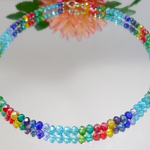 May include: A multi-strand necklace composed of small, faceted beads in a rainbow of colors. The beads transition from light blue to red, yellow, green, and purple. The necklace has a silver-colored clasp and is displayed on a reflective surface.