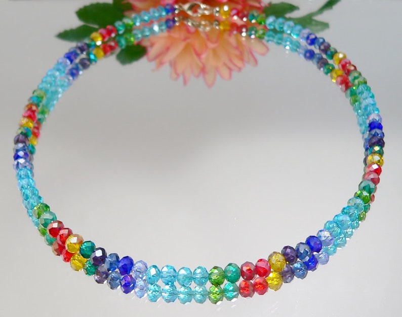 May include: A multi-strand necklace composed of small, faceted beads in a rainbow of colors. The beads transition from light blue to red, yellow, green, and purple. The necklace has a silver-colored clasp and is displayed on a reflective surface.