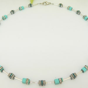 Peut inclure: Un collier argenté avec des perles carrées turquoise et grises. Les perles sont rehaussées de strass transparents.