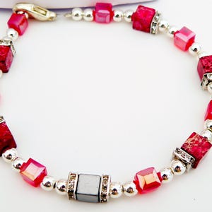 Può includere: Un braccialetto caratterizzato da un motivo ripetitivo di perline rosse a forma di cubo con texture marmorizzata, perline quadrate rosa traslucide più piccole e perline sferiche color argento metallizzato. Una perla cubica grigia è centrata e affiancata.
