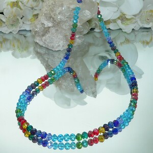 May include: A multi-strand necklace composed of small, faceted beads in a rainbow of colors. The beads transition from deep purple to light blue, with red, yellow, and green accents. The necklace is displayed on a reflective surface.
