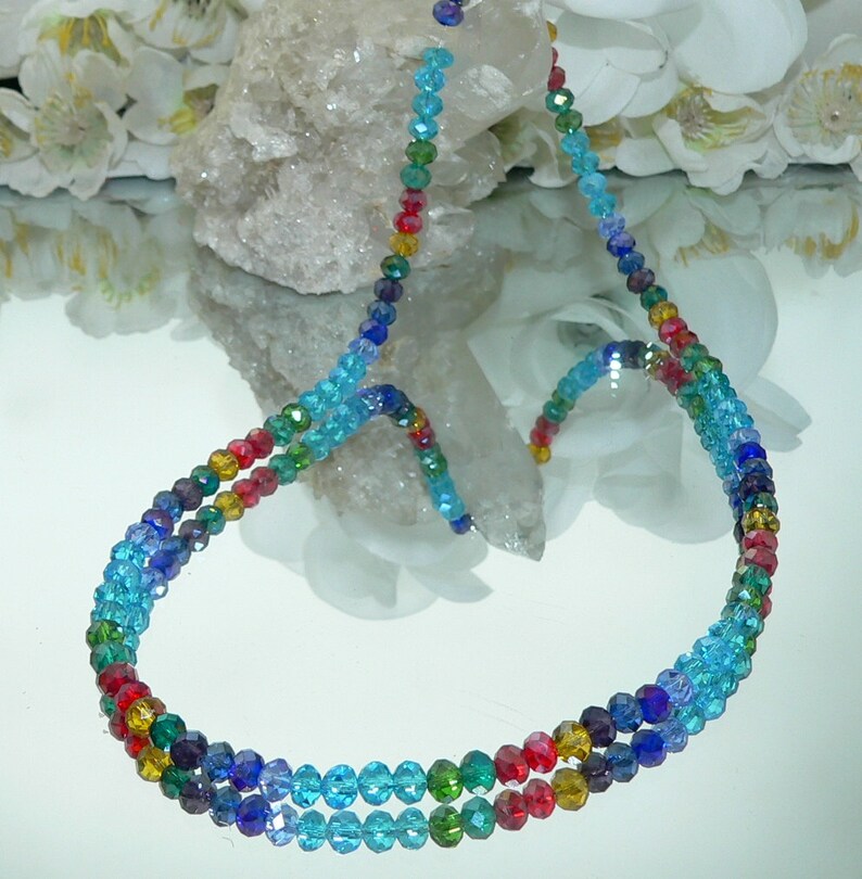 May include: A multi-strand necklace composed of small, faceted beads in a rainbow of colors. The beads transition from deep purple to light blue, with red, yellow, and green accents. The necklace is displayed on a reflective surface.