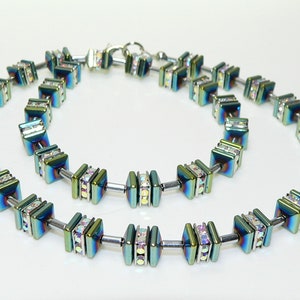 May include: A multi-strand necklace with iridescent green and blue square beads with clear crystal accents. The necklace is made of metal and has a silver finish.