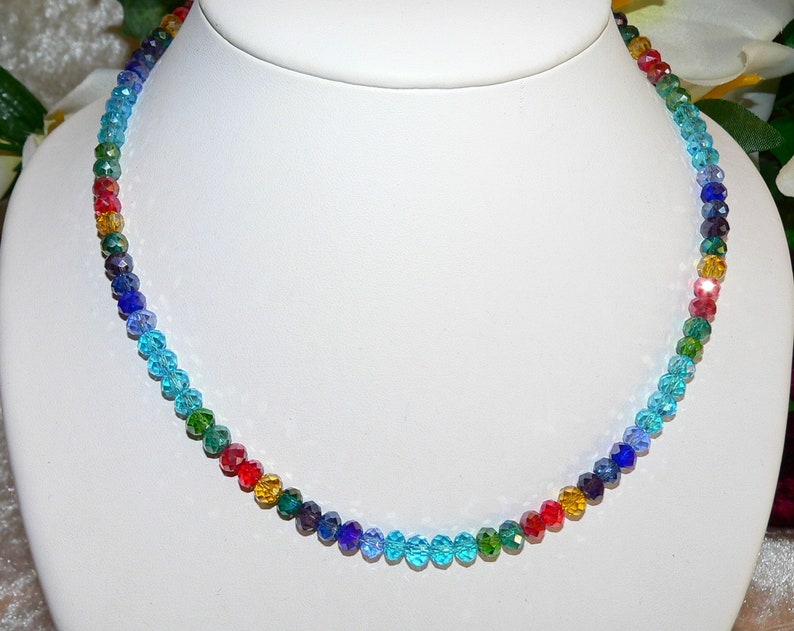 May include: A colorful beaded necklace displayed on a white mannequin. The necklace features a gradient of colors, including red, orange, yellow, green, blue, and purple. The beads are small and faceted, reflecting light.