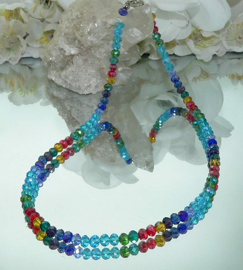 May include: A multi-strand necklace featuring a gradient of colorful beads. The beads transition from red to yellow, green, blue, and purple. The necklace has a silver clasp and is displayed on a reflective surface.
