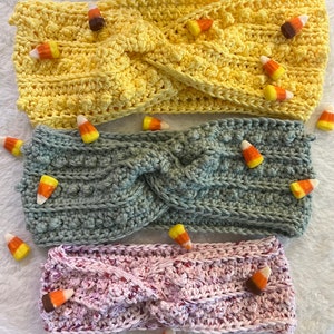 May include: Three handmade crochet headbands in yellow, gray-green, and pink, each adorned with candy corn. The headbands have a twisted design and a textured stitch pattern. Perfect for fall or Halloween.