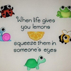 May include: A white cross-stitch with the saying "When life gives you lemons, squeeze them in someone's eyes". The cross-stitch is decorated with colorful felt appliques of a ladybug, a frog, a snail, a bee, and a turtle.