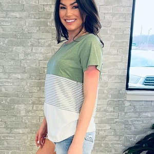May include: A short-sleeve t-shirt featuring a color-blocked design with an olive green top section, a middle section with thin horizontal white and gray stripes, and a solid white bottom section, paired with light blue denim frayed shorts.