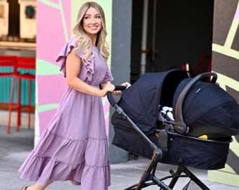 Puede incluir: Una persona con un vestido lavanda empuja un cochecito negro. El cochecito tiene un marco negro y un capazo. La persona sonríe y camina por una acera. Producto para bebés.