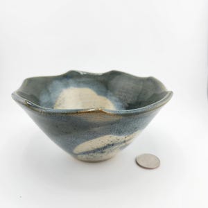 May include: A handmade ceramic bowl with a unique, scalloped rim. The bowl features a blend of blue and off-white glaze, creating a marbled effect. The bowl's diameter is approximately 6 inches.
